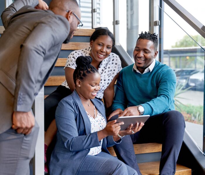 Four professional male and female coworkers sitting on staircase smiling practicing teamwork on corporate tablet